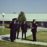 Photograph of six men standing outside, in front of a rectangular building. They each wear a black uniform suit. Each suit has shiny details and a badge on one sleeve. The sun shines onto the paved grass.