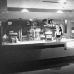 Black and white photo of a snack / food counter. There are different food and drink machines behind the counter.