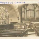Black and white photo inside a church. There are rows of pews and a very large pipe organ behind the altar. To the left is an impressive stained glass window.