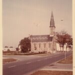 Colour photo of a large church with a tall spire. There is a zig zag pattern on the roof.