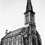 Black and white photo of a large church with a tall spire. There is a zig zag pattern on the roof.