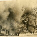 Sepia toned photo of a building on fire, with smoke billowing. There are a number of people in the photo, assessing the scene