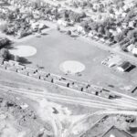 Black and white aerial photo showing a park, rail line, and surrounding trees.