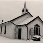 Colour photo of a long white building with two darker triangular roofs and a spire, everything is coated in a thin layer of snow