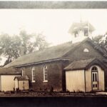 Sepia photo of brick building with two smaller white building attachments, all have darker triangular roofs and the main segment has a spire.
