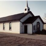 Colour photo of a long white building with darker triangular roofs and a spire.