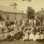 Sepia image of a long school with a spire on top students standing out front.