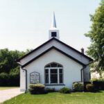 Colour photo of the front of a long white building with darker triangular roofs and a spire.