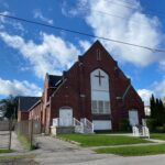 Colour photogrpah of a red brick church with a triangular roof. There is a large cross in the middle of the building facade.