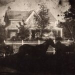 Sepia photo of a stone house, with two men in carriages out front of a white picket fence